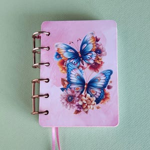 May include: A small pink notebook with a butterfly design. The cover features two blue butterflies with orange and white accents, surrounded by flowers. The notebook has a gold spiral binding and a pink ribbon bookmark with a decorative charm.
