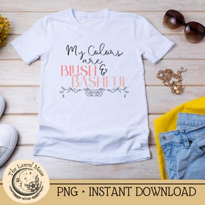 May include: White t-shirt featuring the text "My Colors are Blush & Bashful" in black and pink script, with a floral design. Sunglasses, jewellery, and clothing are placed near the shirt.
