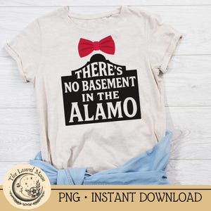 Puede incluir: Camiseta beige con la frase "THERE'S NO BASEMENT IN THE ALAMO" en texto negro sobre un letrero negro, coronada con una pajarita roja. La camiseta está doblada, con una camisa vaquera azul visible.