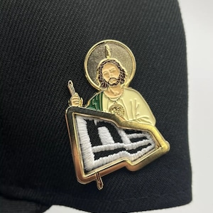 May include: Gold-tone metal pin featuring a depiction of Jesus holding a flag with the New Era logo. The flag is white with black stripes and a green border.