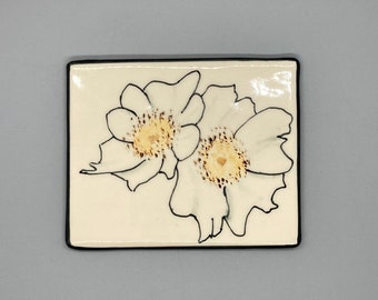 May include: White ceramic tile with a black outline featuring a hand-painted design of two white flowers with brown centers.