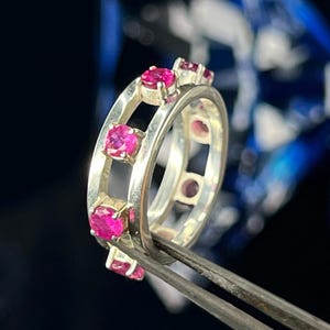 Puede incluir: Un anillo de plata con múltiples piedras preciosas de color rosa brillante. El anillo tiene un diseño abierto con recortes cuadrados y está sujeto por unas pinzas. Las piedras preciosas están espaciadas uniformemente alrededor del anillo.