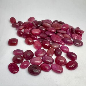 Puede incluir: Una colección de numerosas piedras preciosas sueltas, pulidas y de forma ovalada en varios tonos de rosa intenso, magenta y rojo oscuro. Cada piedra presenta una superficie lisa y redondeada con un acabado brillante, mostrando variaciones naturales.