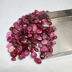 Puede incluir: Una colección de piedras preciosas pulidas sueltas en varios tonos de rosa, magenta y rojo intenso. Las piedras son de forma ovalada, redonda e irregular, con superficies lisas y reflectantes. Una pala de metal está parcialmente enterrada en el montón.