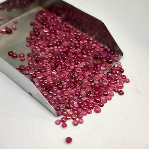 Puede incluir: Una pala de metal llena de una gran colección de pequeñas piedras preciosas redondas, translúcidas, de color rosa y rojo intenso. Las piedras varían en tono e intensidad, mostrando una textura de superficie lisa y pulida al derramarse.