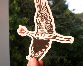Peut inclure: Un autocollant représentant une illustration stylisée d'un oiseau en vol. L'oiseau est représenté en noir et blanc avec un détail en forme d'éclat d'étoile rouge. L'autocollant est tenu devant un fond de feuillage vert.