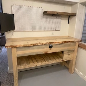 May include: A light-colored wooden desk with a live edge top and a drawer. The desk has a lower shelf and is supported by four sturdy legs. A pegboard and shelf are mounted on the wall behind the desk.