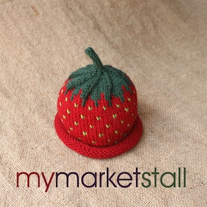 May include: A knitted strawberry hat with a green top and red body. The hat is on a brown burlap background.