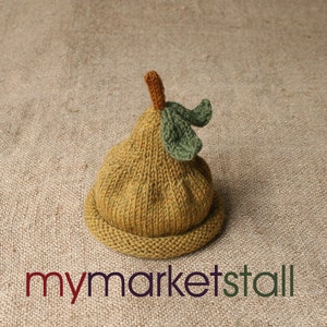 May include: A knitted pear-shaped hat for a baby, in a light brown color with green leaves on top. The hat is sitting on a burlap background.