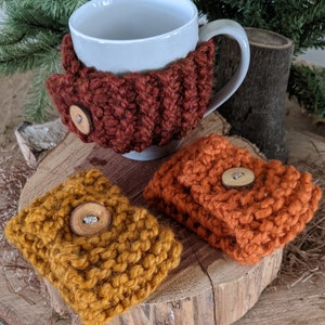 May include: A white ceramic mug with a rust-colored knitted cozy, and two additional cozies in mustard yellow and orange. Each cozy has a wooden button closure. The items are displayed on a wooden surface with a natural background.