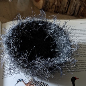 May include: A black felt nest with a fluffy white rim. The nest is empty and sits on a book.