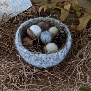 May include: A small, gray felt bowl filled with decorative acorns, pinecones, and felt balls in shades of white and gray. The bowl sits on a bed of brown paper and is surrounded by dried leaves, creating a rustic, autumnal theme.