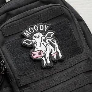 May include: Black backpack with a patch featuring a cartoon cow with the word "MOODY" above it. The cow is white with black spots and a pink udder. The backpack has multiple compartments and straps.