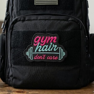 May include: Black backpack with a patch that reads "gym hair don't care" in pink and teal. A rolled-up brown towel, a water bottle, and keys are next to the backpack. The backpack has an American flag patch.