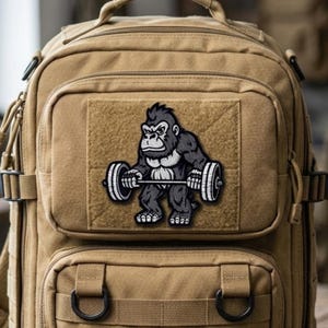 May include: Tan tactical backpack with multiple compartments and straps. A patch featuring a black and gray gorilla lifting a barbell is affixed to the front pocket. The gorilla patch is a bold design element.