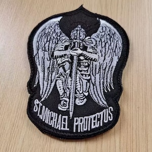 May include: Black and white embroidered patch featuring a winged angel holding a sword. The text "St. Michael Protectus" is embroidered below the angel.