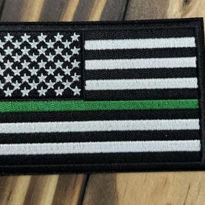 May include: A rectangular embroidered patch featuring a stylized American flag design. The patch consists of black and white horizontal stripes with a field of white stars on a black background, accented by a single horizontal green stripe.
