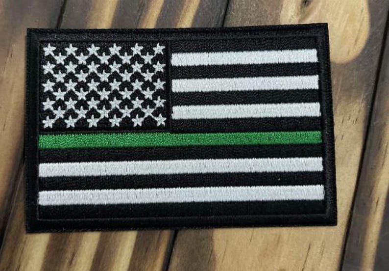 May include: A rectangular embroidered patch featuring a stylized American flag design. The patch consists of black and white horizontal stripes with a field of white stars on a black background, accented by a single horizontal green stripe.