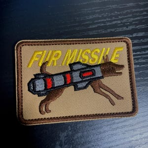 May include: A tan embroidered patch with a brown dog carrying a gray missile with red stripes. The text "FUR MISSILE" is embroidered in yellow above the dog.