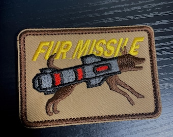 May include: A tan embroidered patch featuring a brown dog carrying a grey missile with red stripes. The text "FUR MISSILE" is embroidered in yellow above the dog.