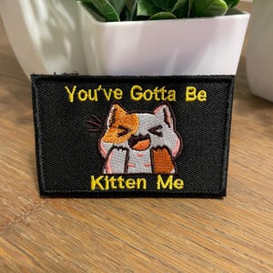 May include: Black embroidered patch with a white cat with orange and brown markings. The cat is laughing and the text "You've Gotta Be Kitten Me" is embroidered in yellow.