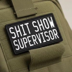 May include: Olive green tactical backpack with a black and white patch that reads "SHIT SHOW SUPERVISOR". The patch is rectangular and features bold, white capital letters. The backpack has multiple straps and a textured surface.