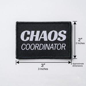 Pode incluir: Um patch retangular preto com texto bordado branco que diz "CHAOS COORDINATOR". O patch tem aproximadamente 7,6 cm de largura e 5,1 cm de altura.
