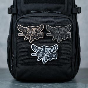 May include: Three embroidered patches with a military emblem featuring a skull, wings, and a weapon. The patches are in shades of tan, gray, and white, and are attached to a black backpack. The patches are detailed and have a tactical theme.