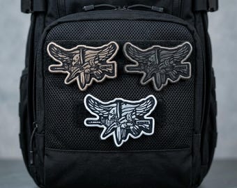 Pode incluir: Três patches bordados com um emblema militar com uma caveira, asas e uma arma. Os patches são em tons de bege, cinza e branco e estão presos a uma mochila preta. Os patches são detalhados e têm um tema tático.