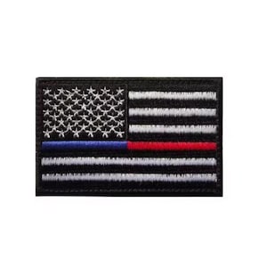 May include: A rectangular embroidered patch featuring a stylized American flag design. The flag consists of black and white horizontal stripes with a field of white stars on a black background, accented by a single horizontal blue and red line.