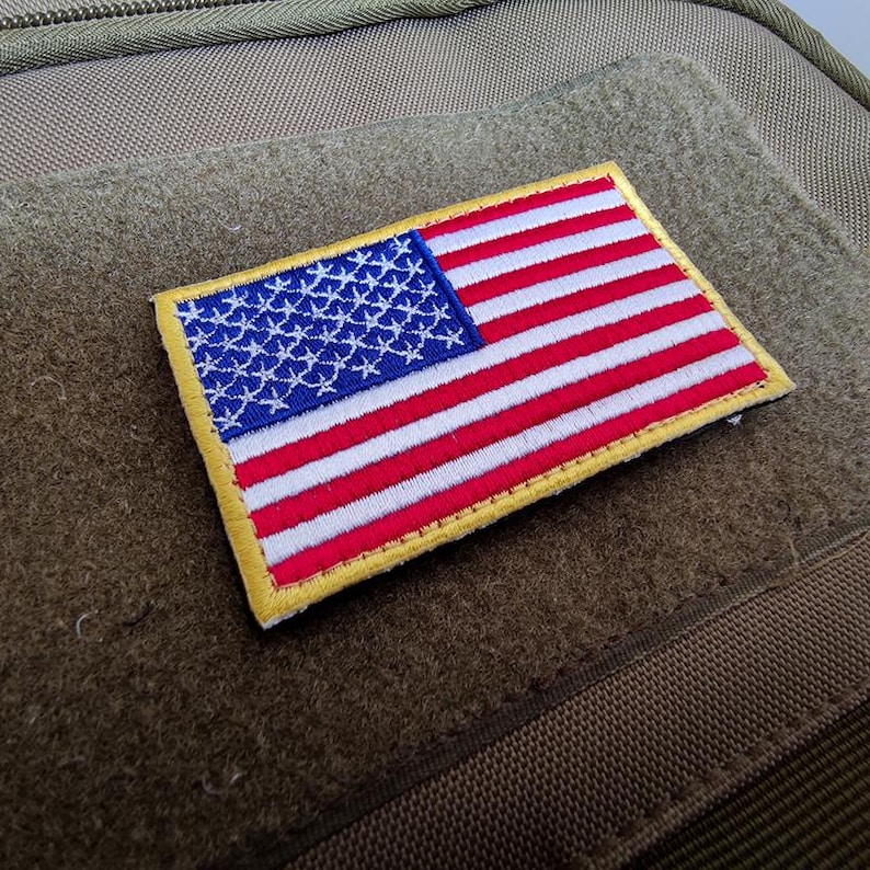 May include: A rectangular embroidered patch featuring the United States flag with a blue canton containing white stars and alternating red and white horizontal stripes. The patch has a thick yellow border and is attached to a tan fabric surface.