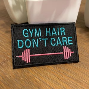 May include: Black rectangular patch with the text "GYM HAIR DON'T CARE" in turquoise, and a pink barbell design. The patch is likely for clothing or accessories, and is a humorous gym-themed item.