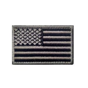 May include: A rectangular embroidered patch featuring a monochromatic American flag design. The patch displays a field of stars on the left and horizontal stripes on the right, rendered in alternating shades of black and dark grey with a stitched border.
