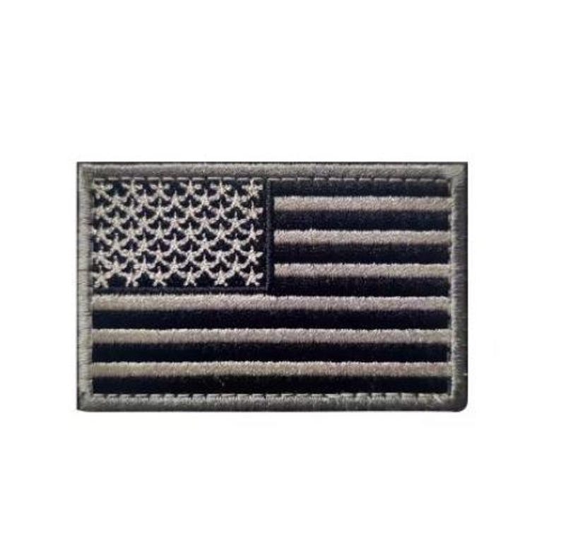 May include: A rectangular embroidered patch featuring a monochromatic American flag design. The patch displays a field of stars on the left and horizontal stripes on the right, rendered in alternating shades of black and dark grey with a stitched border.