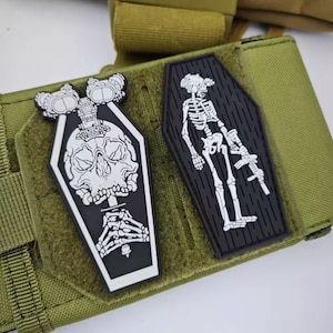 May include: Two black and white embroidered patches with a skeleton and skull design. The patches are attached to a green tactical vest.