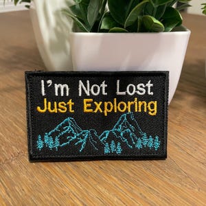 May include: Black embroidered patch with white text that reads "I'm Not Lost Just Exploring" and a blue mountain range with green trees.