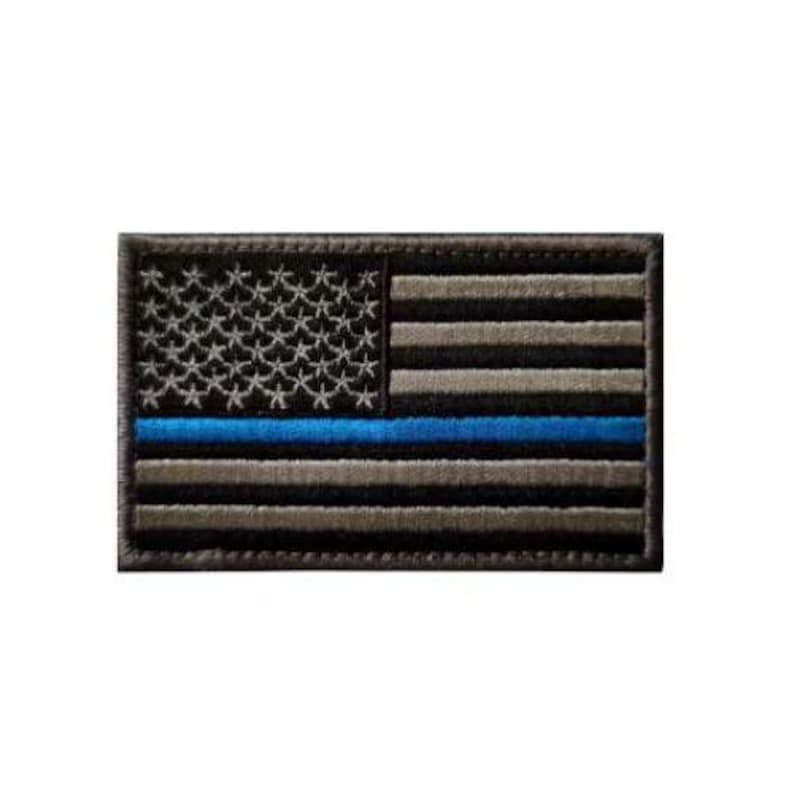 May include: A rectangular embroidered patch featuring a stylized American flag design. The flag consists of black and grey horizontal stripes with a single horizontal blue stripe, and a black field with grey embroidered stars.