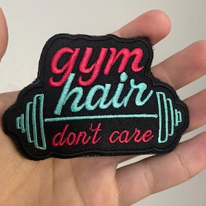 May include: Black embroidered patch with the text "gym hair don't care" in red and teal. The patch features a barbell design and is likely for a gym bag or clothing.