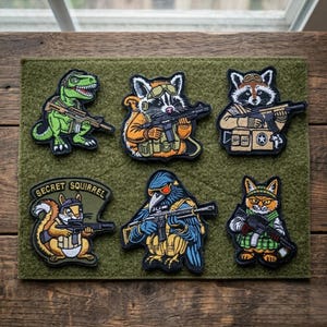May include: A collection of six embroidered patches featuring various animals, including a dinosaur, raccoon, squirrel, and cat, each holding a rifle. The patches are in vibrant colors and set on a green background. The squirrel patch has the text "SECRET SQUIRREL".