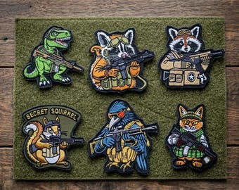 Pode incluir: Uma coleção de seis patches bordados com vários animais, incluindo um dinossauro, um guaxinim e um esquilo, cada um segurando um rifle. Os patches são em cores vibrantes e colocados em um fundo verde. O patch do esquilo tem o texto "SECRET SQUIRREL".