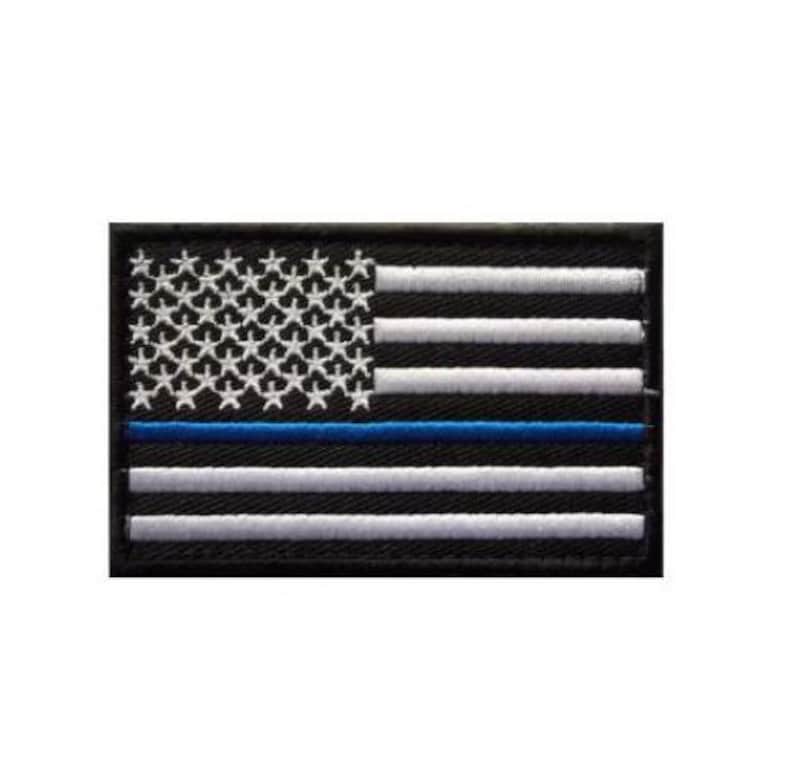 May include: A rectangular embroidered patch featuring a stylized American flag design. The flag is rendered in black with white embroidered stars and horizontal stripes, accented by a single horizontal blue stripe across the center.