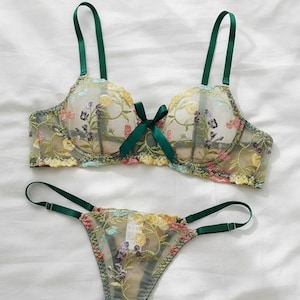 May include: A delicate lingerie set with floral embroidery. The bra and thong feature colourful flower patterns and green detailing, including straps and a bow. The set is presented on a white background.