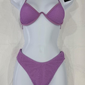 May include: A two-piece lilac bikini set displayed on a white mannequin. The bikini top has a halter neck design and the bottoms are high-cut. The fabric has a textured appearance. A tag is attached to the top.