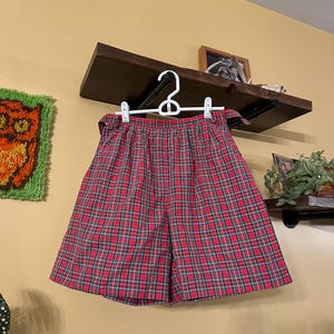 May include: Red plaid shorts hanging on a white hanger. The shorts have a classic plaid pattern in red, black, and white. The shorts are made of a lightweight fabric and have a high waist. The shorts are displayed against a yellow wall.
