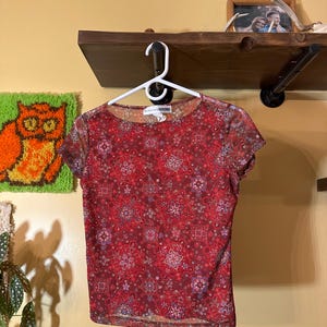 May include: A short-sleeved, sheer red top with a floral pattern. The top is hanging on a white hanger. The floral design includes shades of red, pink, and purple. The top is made of a lightweight material.