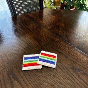 May include: Two rainbow-colored coasters on a dark wood table. The coasters are made of white fabric with a rainbow pattern stitched on.