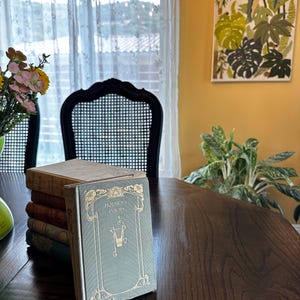 May include: A stack of vintage books with a green cover and gold lettering that reads "Holmes's Poems" on a wooden table. The book is in a room with a window and a chair.