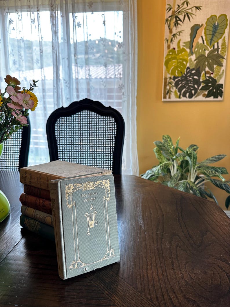 May include: A stack of vintage books with a green cover and gold lettering that reads "Holmes's Poems" on a wooden table. The book is in a room with a window and a chair.