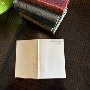 May include: A stack of vintage books with ornate covers, the top book is red with gold detailing and the bottom book is green. The top book has a handwritten inscription on the inside cover that reads "B.F. Gaynor 1/24/174".