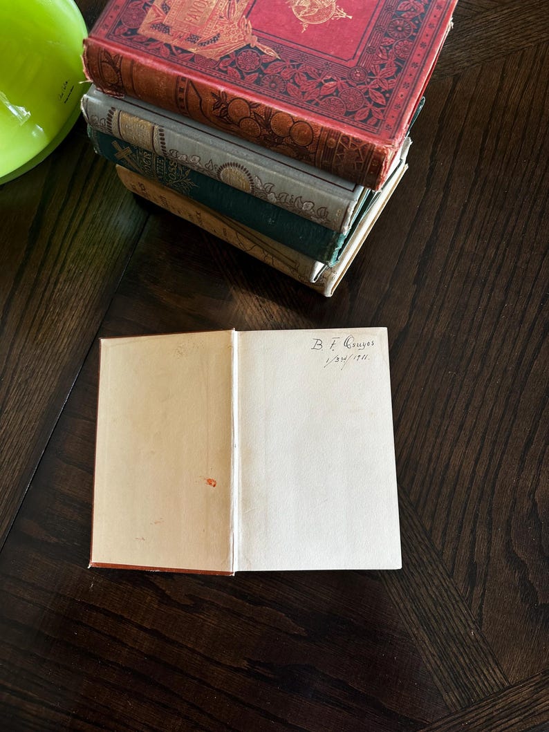 May include: A stack of vintage books with ornate covers, the top book is red with gold detailing and the bottom book is green. The top book has a handwritten inscription on the inside cover that reads "B.F. Gaynor 1/24/174".